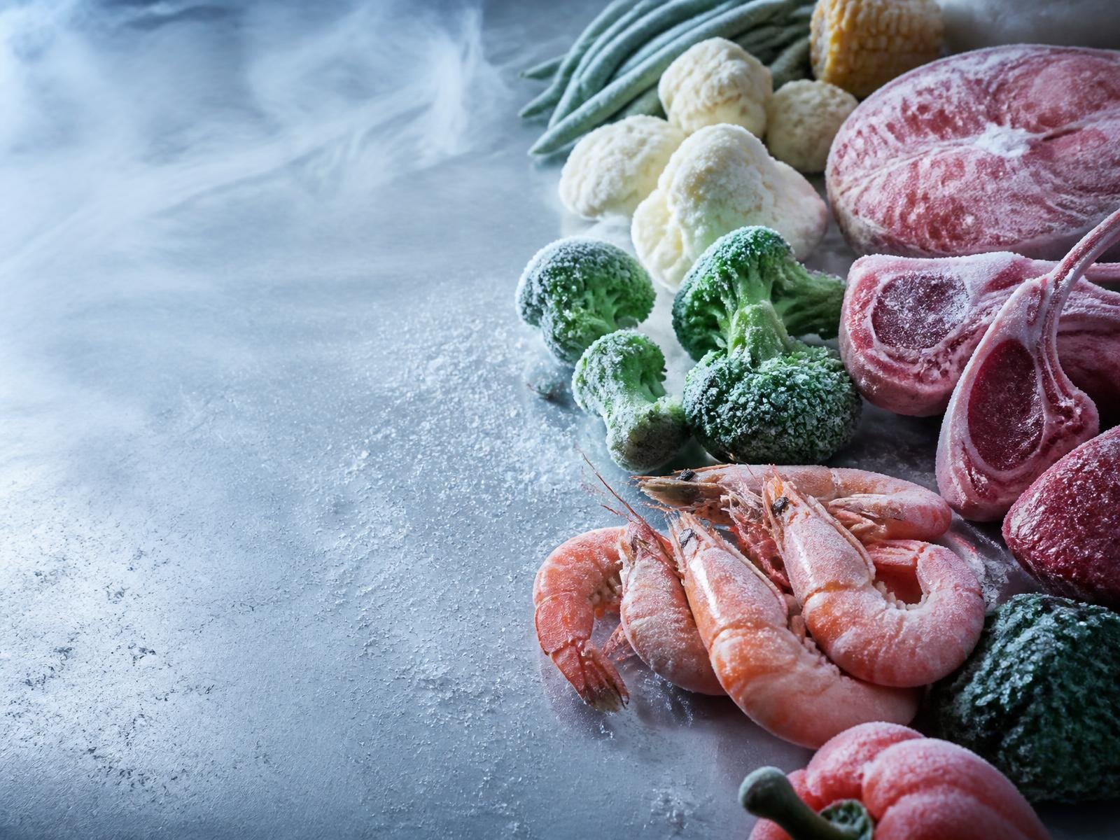 Frost-covered frozen vegetables, prawns and meat cuts on a cold steel surface